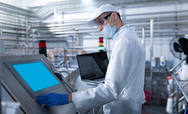 man in a white robe and a mask with a laptop in his hands stands near the digital screen at the factory